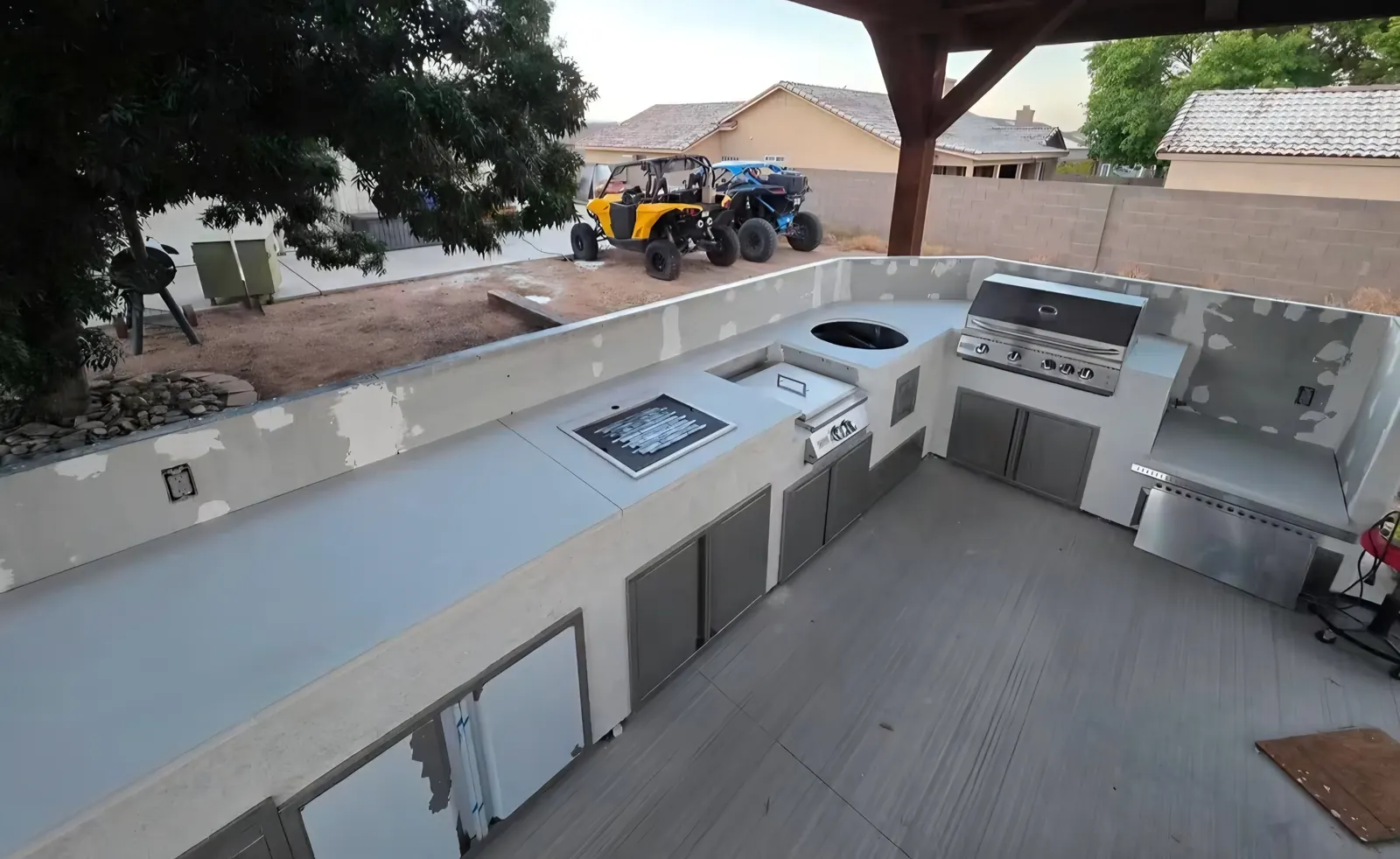 L-shaped outdoor kitchen with grey concrete countertops, built-in grill and stainless doors under a patio cover